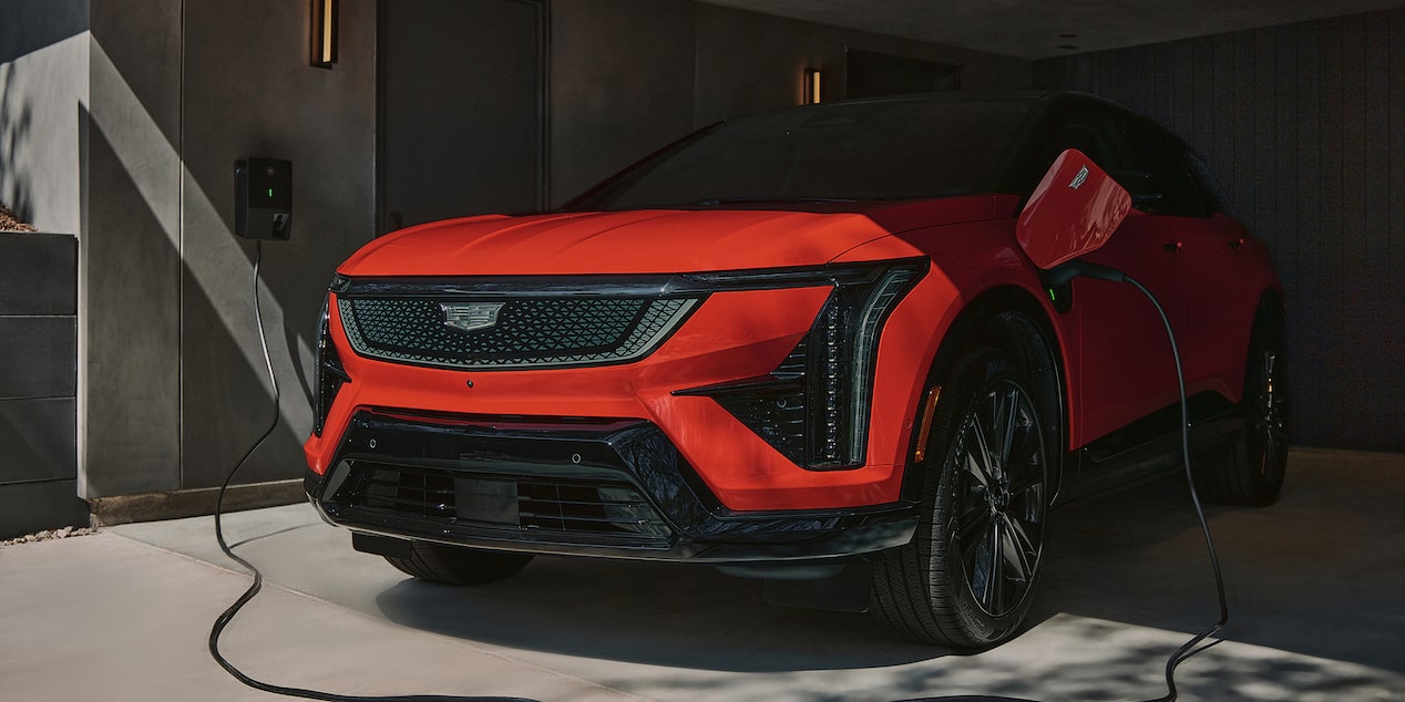 Cadillac OPTIQ 2027 en color Radiant Red circulando por una avenida Cadillac OPTIQ 2027 conectado a una estación de carga eléctrica